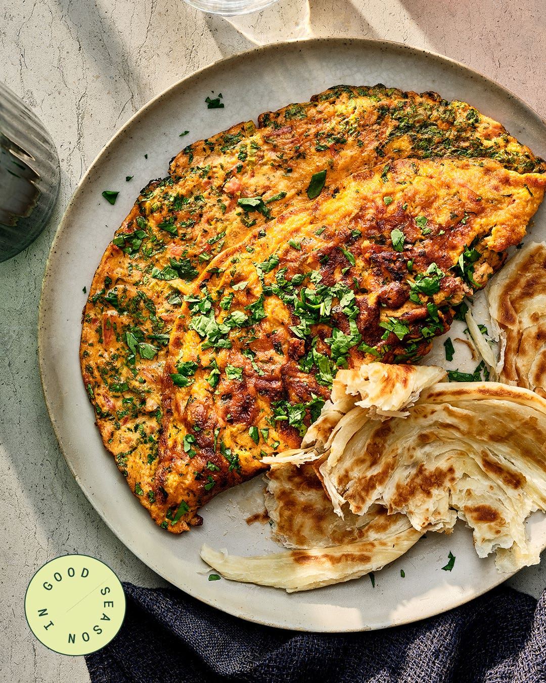 Desi omelet with tomato and scallions, browned and sprinkled with chopped herbs, served on a plate with torn pieces of flaky paratha.