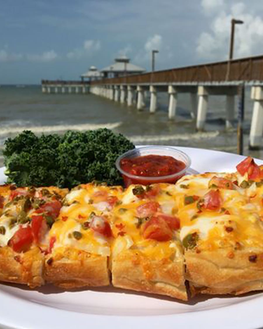 Cheese-covered seasoned bread slices with diced tomatoes, served with a cup of marinara and a leafy green garnish, with a beach and long pier in the background.