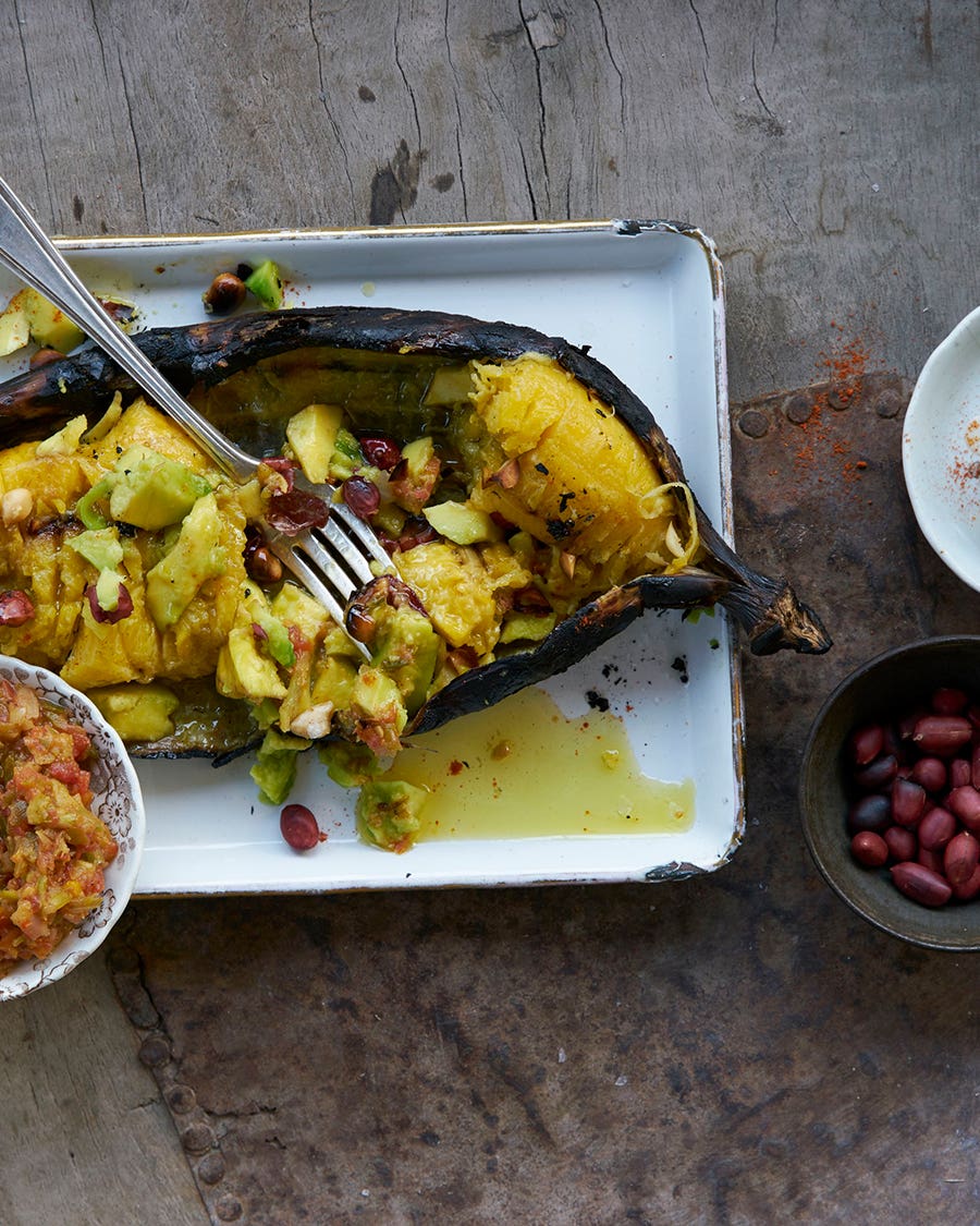 Roasted plantain split open on a tray and topped with diced avocado and peanuts, with a fork on top; bowls of pepper compote, peanuts, and a halved lime alongside.